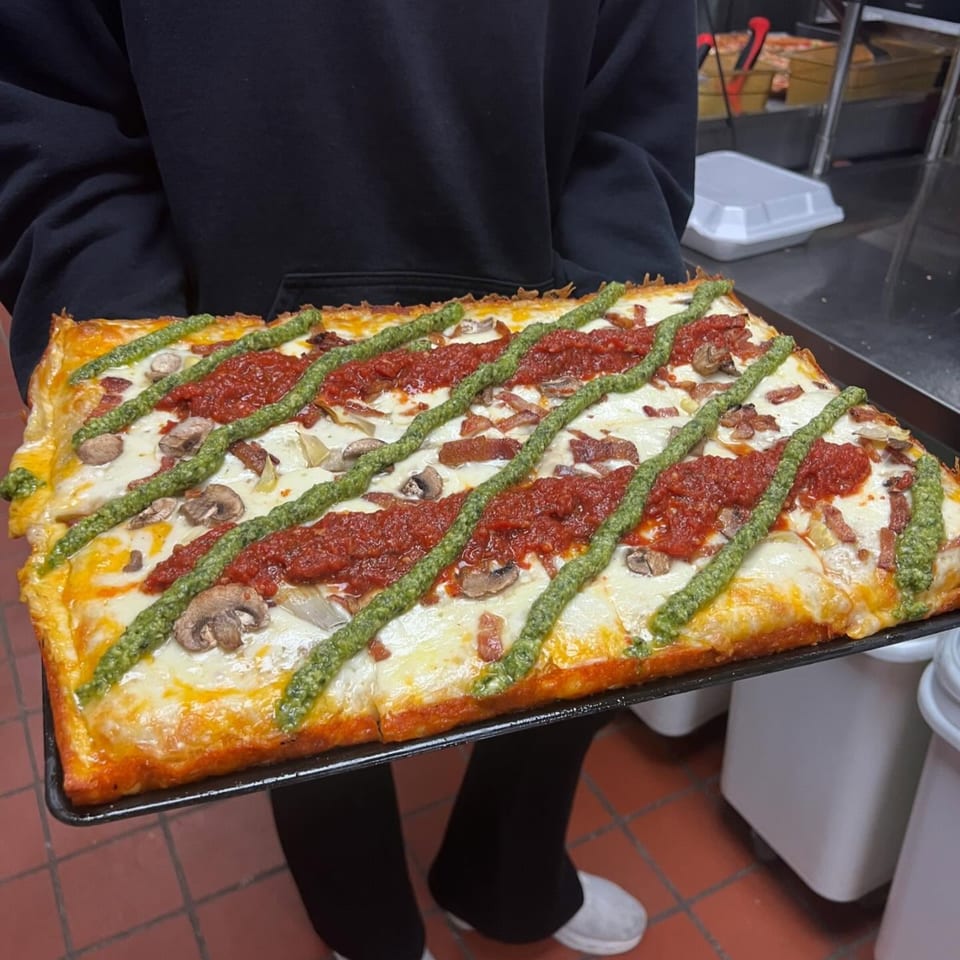 A rectangular TuscNY Pizza with red sauce, pesto, mushrooms, and bacon, held by a person.