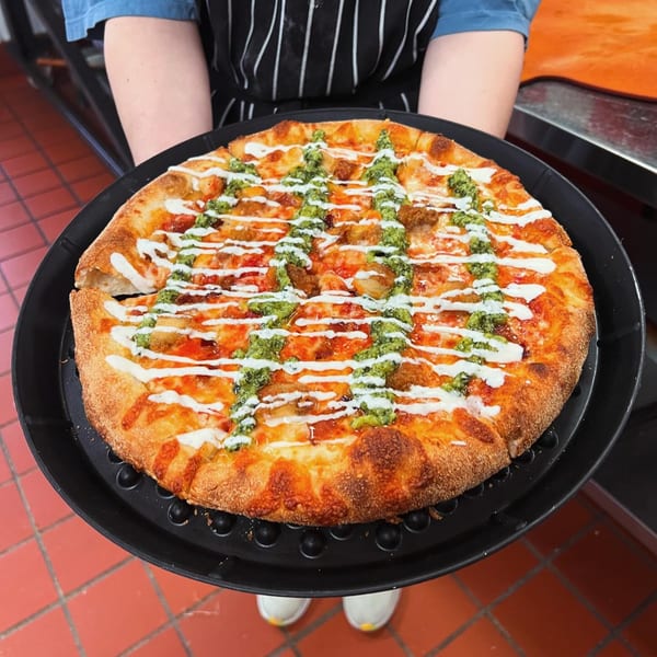 A TuscNY Pizza employee holding a freshly baked pizza topped with pesto, white sauce, and chicken.
