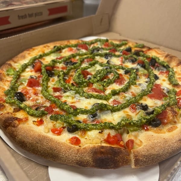 A TuscNY Pizza with pesto swirls, tomatoes, and black olives in a pizza box.
