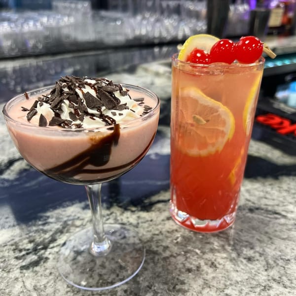 Two cocktails on a bar: a pink creamy drink with chocolate shavings and a red drink with lemon and cherries.