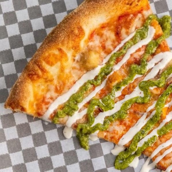 A slice of TuscNY Pizza with melted cheese, green pesto, and white sauce on checkered paper.